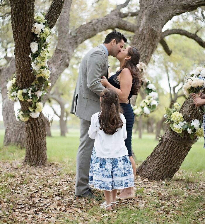 A couple kissing under trees with their adopted daughter watching, representing heartwarming adoption stories.