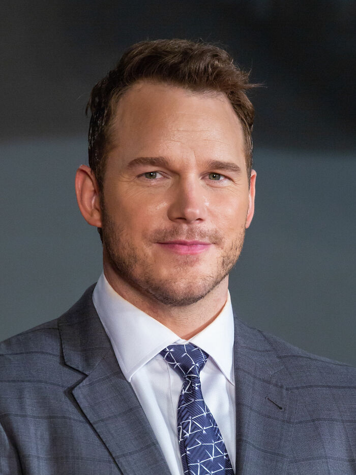 Chris Pratt smiling in a gray suit and patterned tie, showcasing his bio and career highlights.