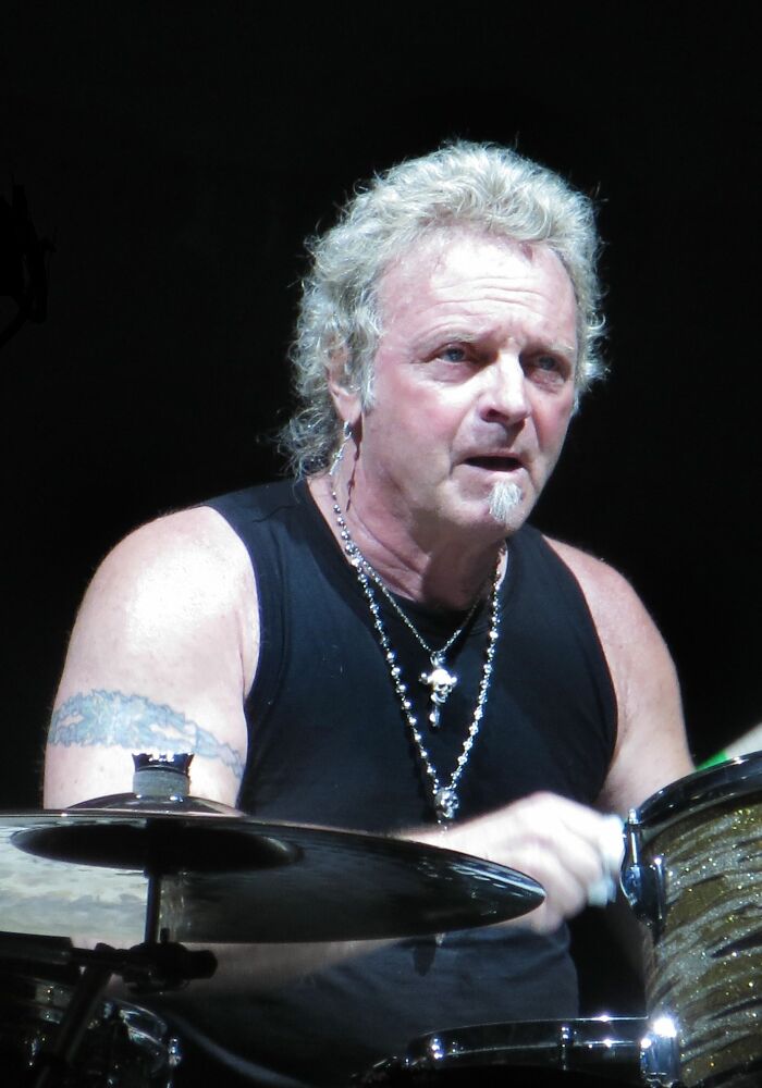 Joey Kramer behind his drum kit, showcasing his impressive bio and career highlights. He has a tattoo on his left arm and two silver necklaces.