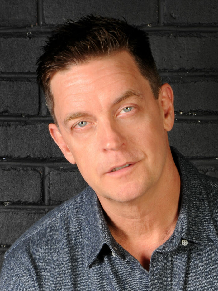 Jim Breuer, comedian, poses for a headshot against a dark brick wall, highlighting his career highlights.