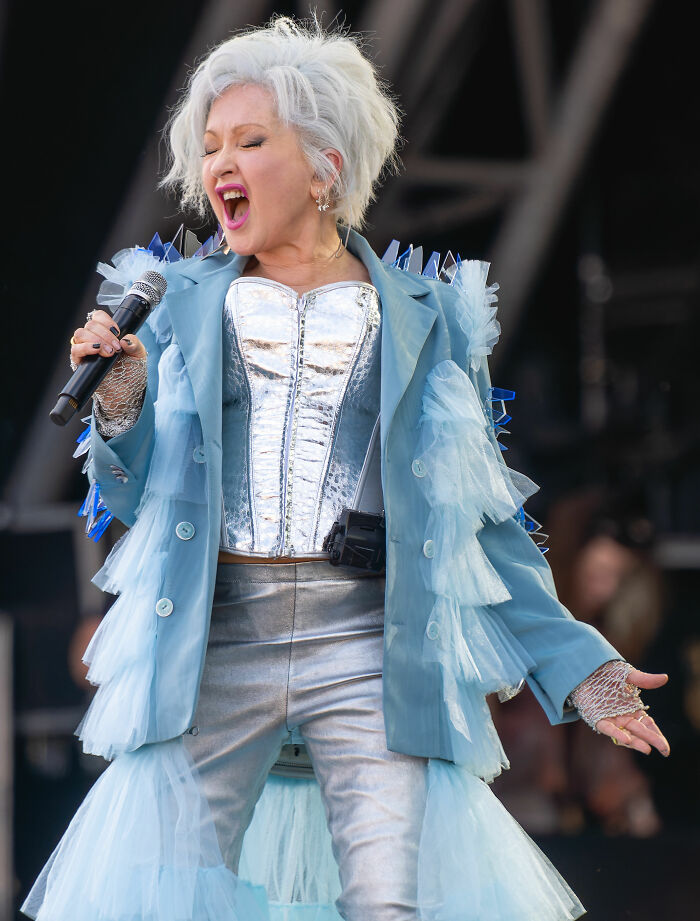 Cyndi Lauper performing live, showcasing her vibrant career highlights. She wears a blue ruffled jacket and silver corset, holding a microphone.