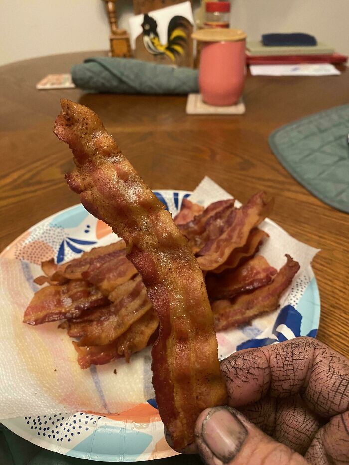 A hand with dirty fingers holding a piece of bacon, with a plate of bacon in the background. Things that should really be left alone.
