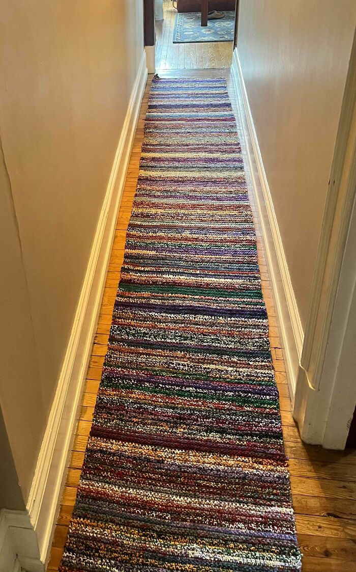 A long, colorful, striped incredible crochet rug stretches down a wooden hallway, handcrafted by a crochet enthusiast.
