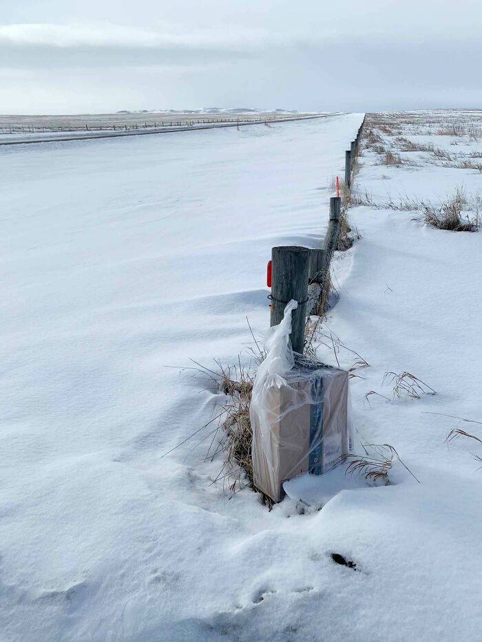 A package left on a snowy fence post next to a road, depicting an unusual delivery fail.