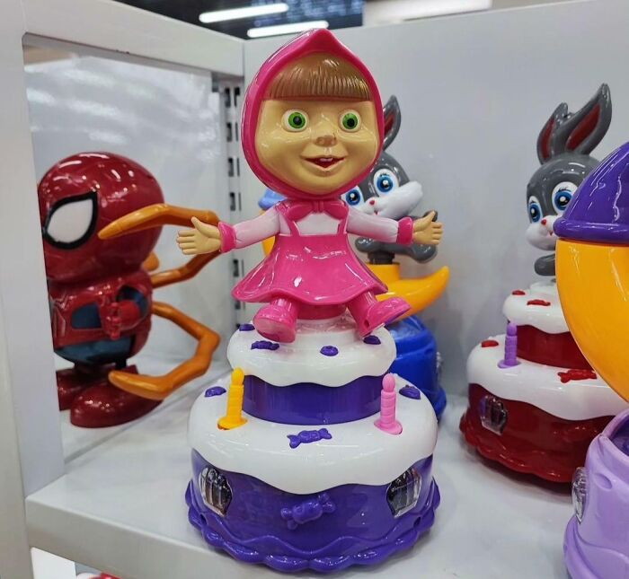 A Masha doll on a birthday cake with a Spiderman figure in the background, a creepy design fail.