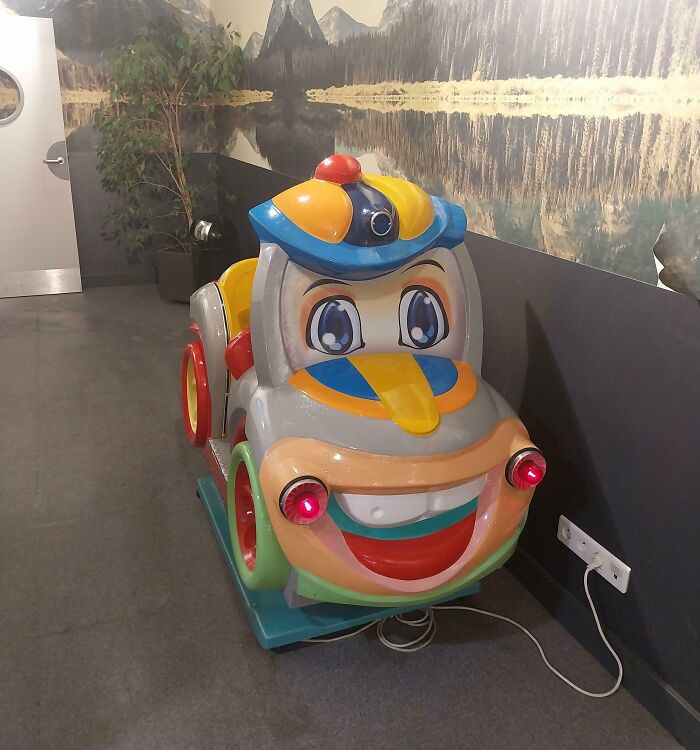 A creepy design fail coin-operated car ride with wide eyes and a big smile, next to a wall with a mountain mural.