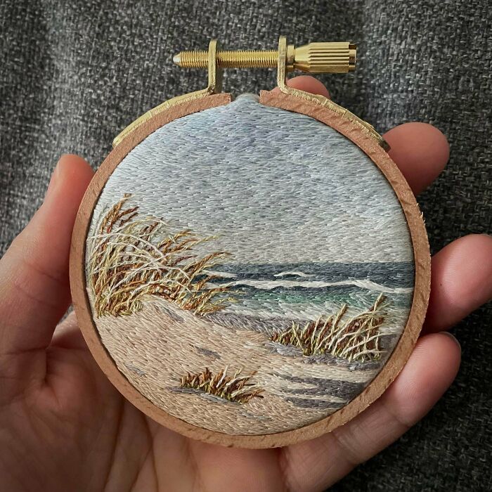 A hand holding a small embroidery hoop showing a serene beach and sand dune scene.