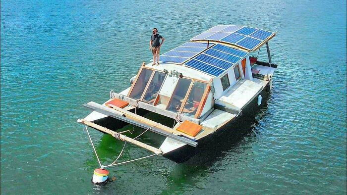A man stands on the roof of a wacky houseboat equipped with solar panels, floating on calm blue water.