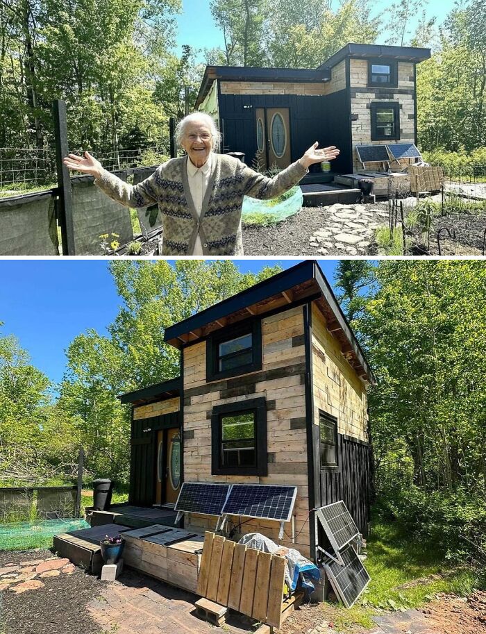 An elderly woman smiling in front of a cool, wacky, and unusual tiny home with solar panels in a lush green setting.