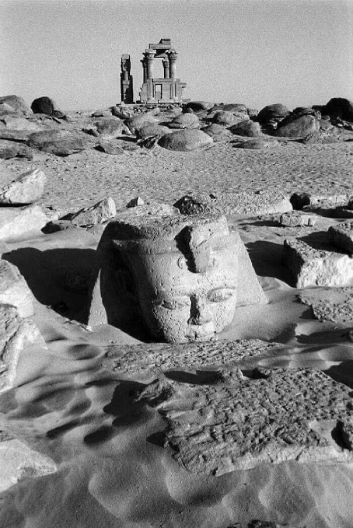 The ancient head of a sphinx partially buried in desert sand with ruins in the background, a very interesting photo.