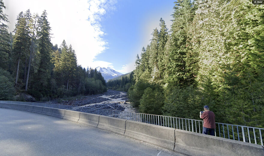 A scenic Google Street View shot of a person taking a picture of a river and mountains surrounded by trees.
