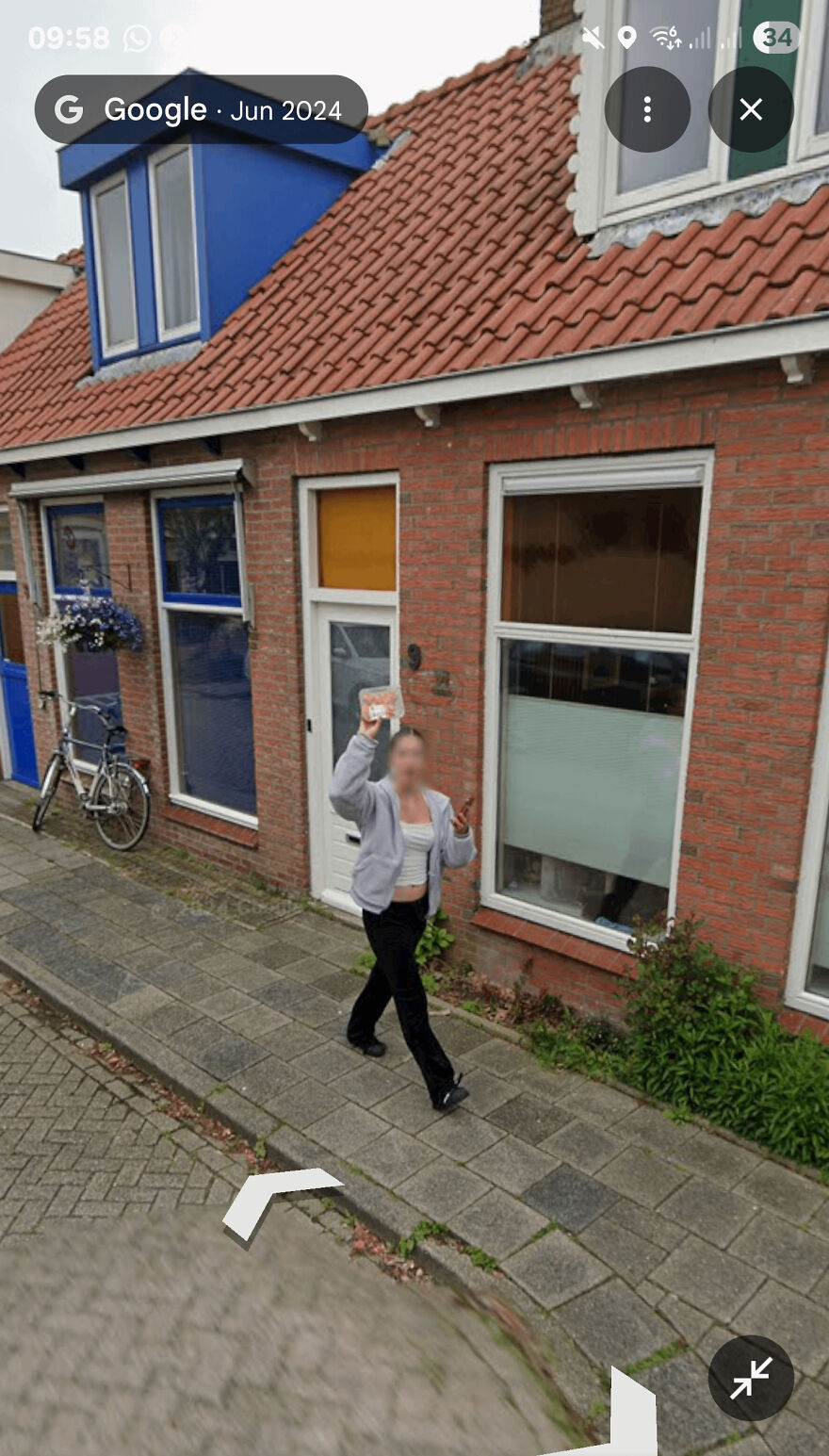 A funny Google Street View photo of a person walking on a sidewalk in front of a brick house, waving.