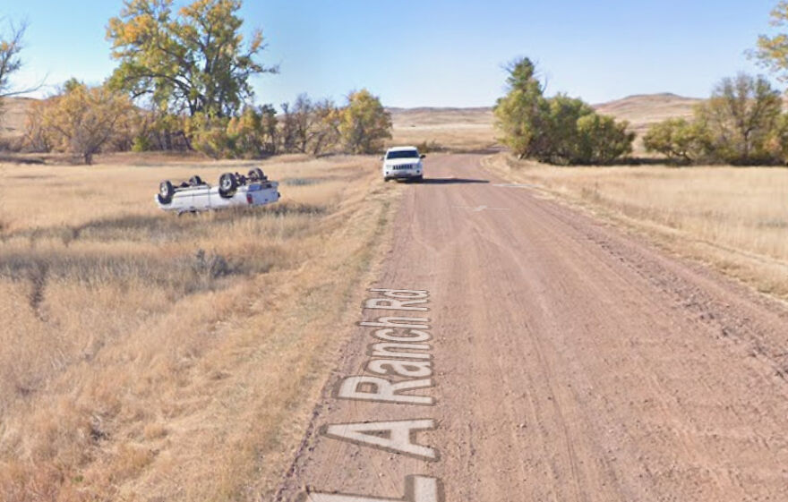 An overturned white truck lies in a dry, grassy ditch next to a dirt road, captured in Google Street View.