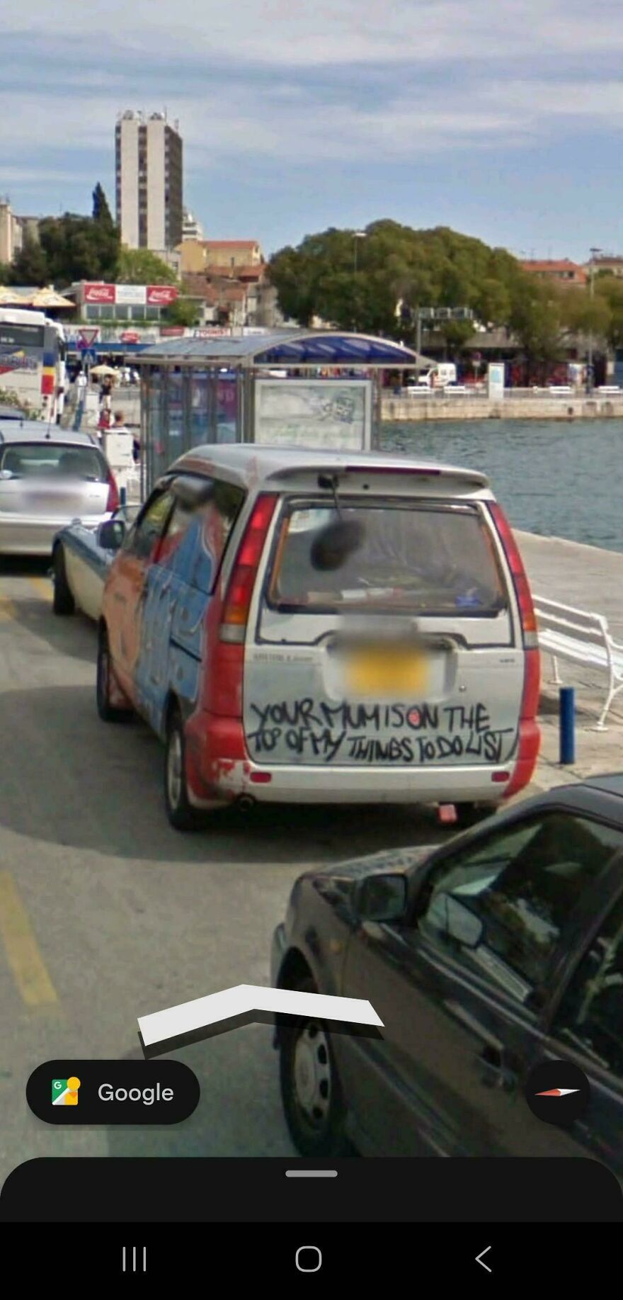 A car with graffiti on its back window parked by the water, seen in a funny Google Street View photo.