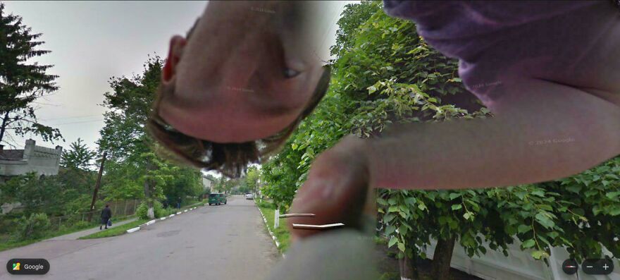 A distorted, close-up human face and arm are captured in a bizarre Google Street View photo on a tree-lined street.