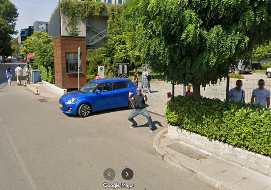 A man in a mask making a funny pose next to a blue car, captured in a Google Street View photo.