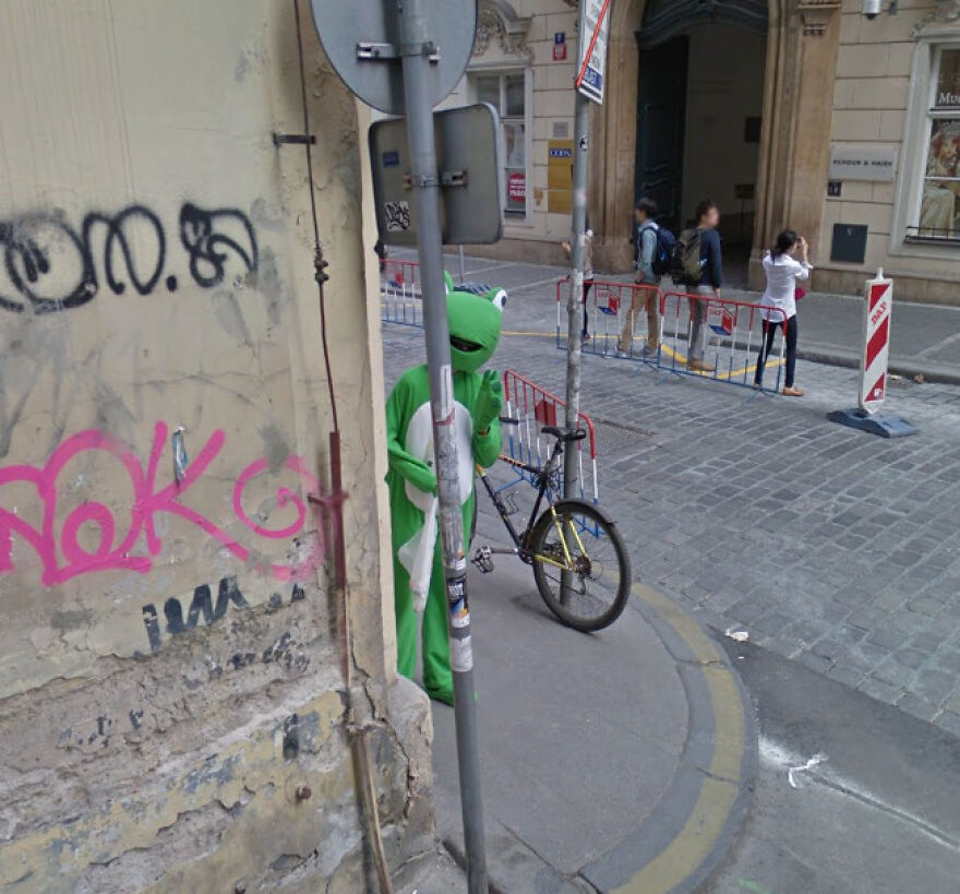 A person in a frog costume stands next to a bicycle on a street with graffiti, a bizarre Google Street View photo.