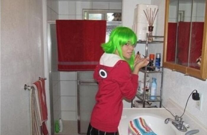 A girl in a green wig applying makeup in a bathroom, with a dog peering from a window, a classic When You See It picture.