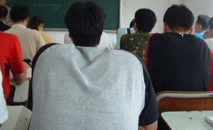 A classroom scene viewed from behind students, one of whom has a hidden face on the back of his head, a clever when you see it picture.