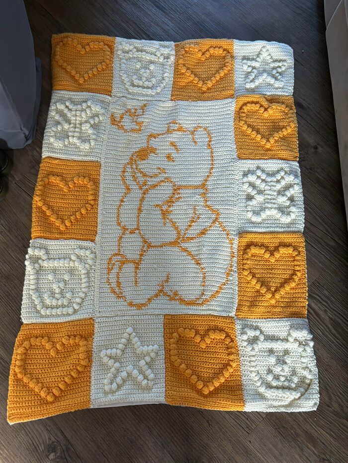 A crochet blanket with a Winnie the Pooh design in the center, surrounded by heart, star, and bear shaped squares, an incredible crochet piece.