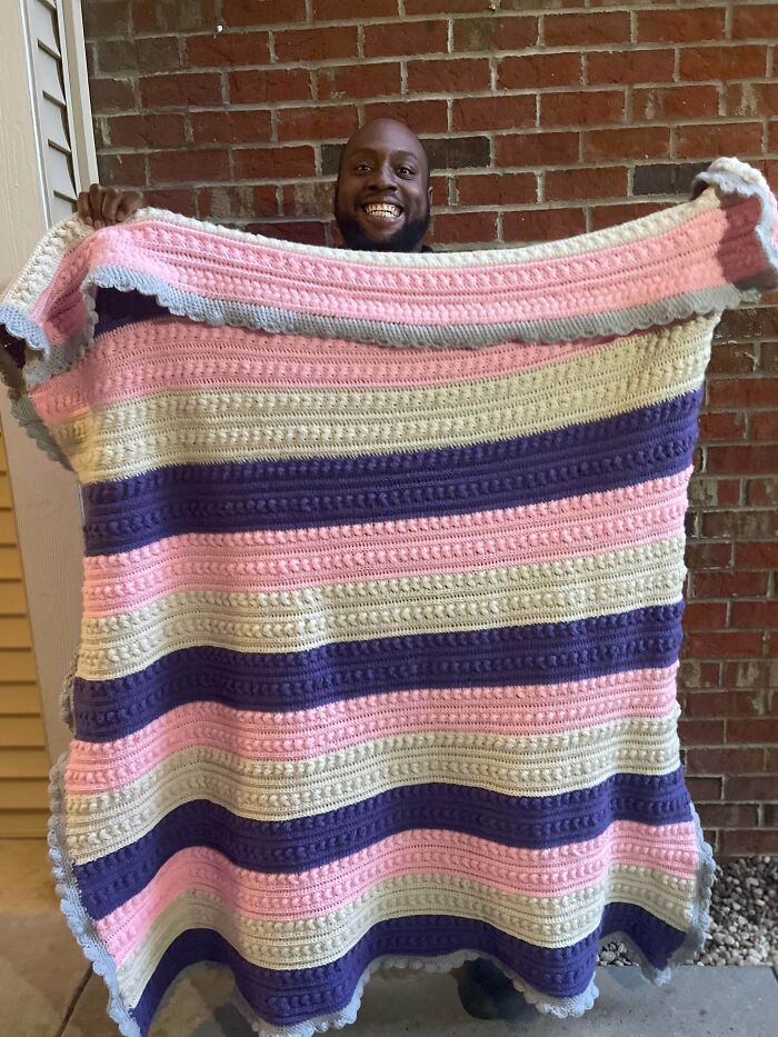 A smiling man holding up a large, colorful crochet blanket with pink, purple, and cream stripes, an incredible crochet item.