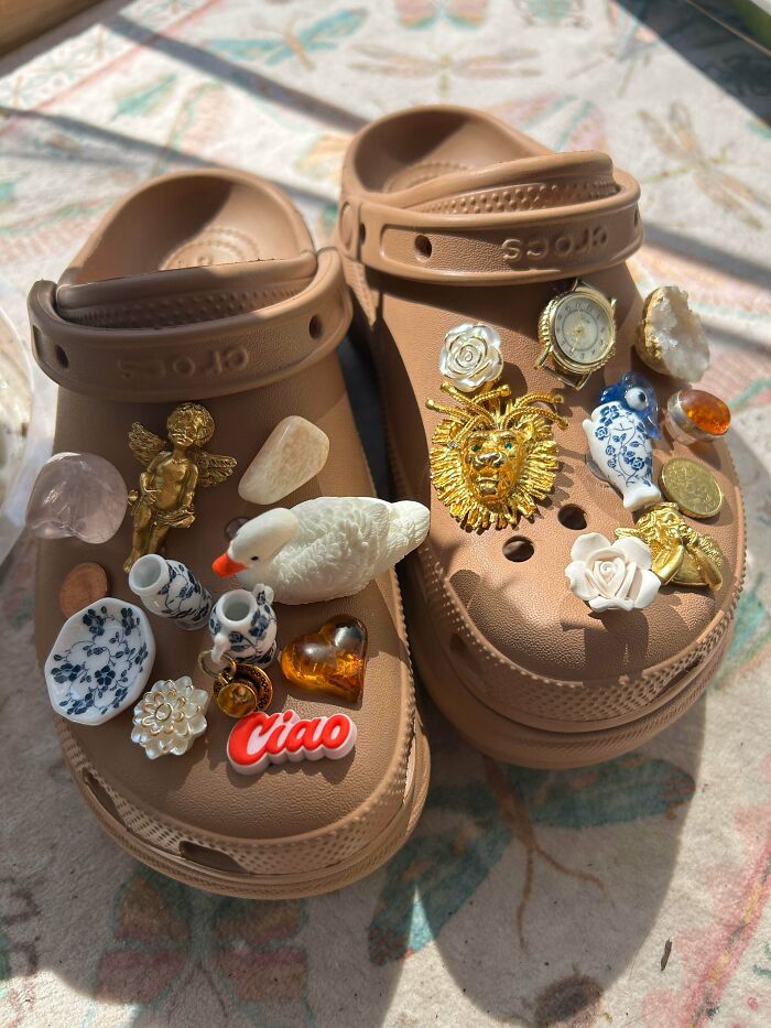 A pair of brown Crocs adorned with various charms and trinkets, made out of things that would’ve been thrown away.
