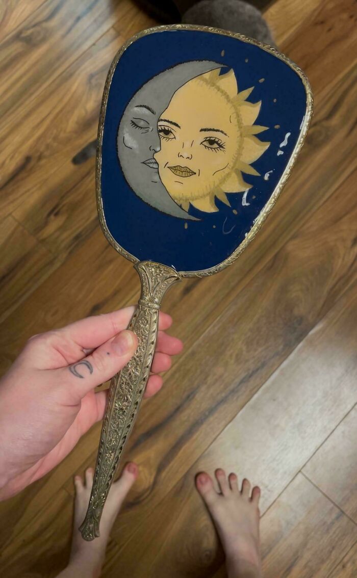 A hand holding a vintage hand mirror with a sun and moon design, made out of things that would’ve been thrown away.