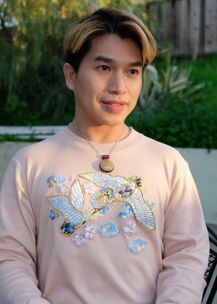 A man wearing a light pink sweatshirt with embroidered cranes and clouds, showcasing his creative style of things made from thrown away items.