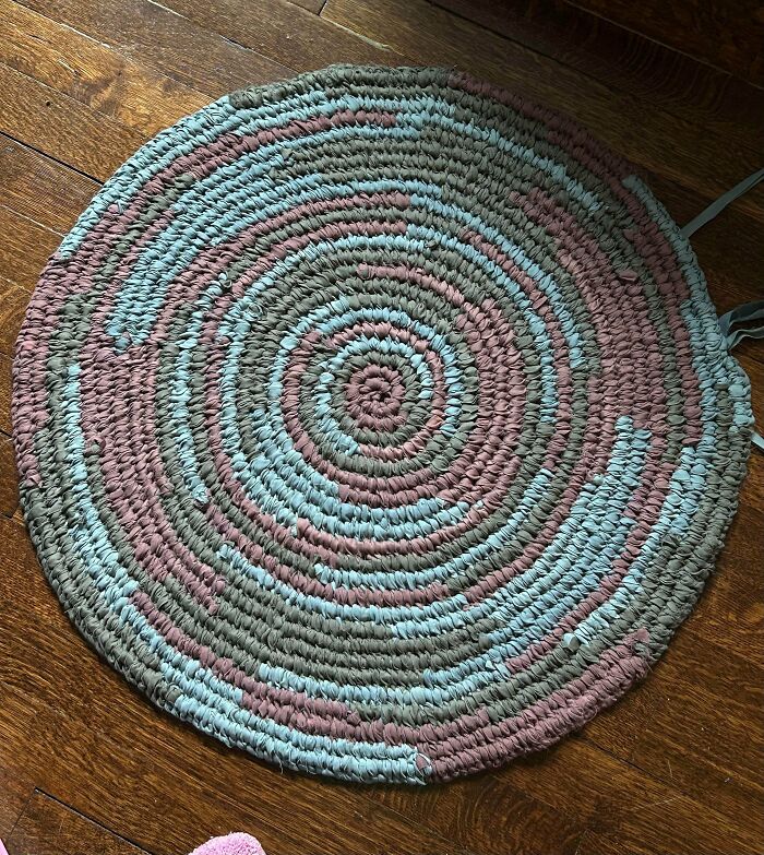 A round braided rug in grey, blue, and pink, made out of things that would've been thrown away on a wooden floor.