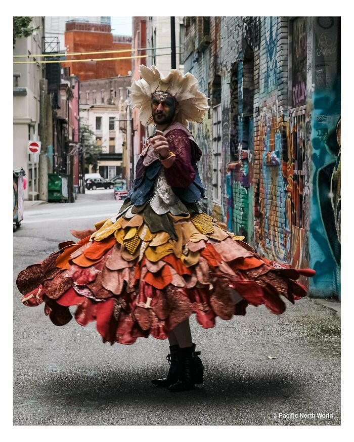 A person in an elaborate, feathered costume made from things that would have been thrown away, dancing in an alleyway.