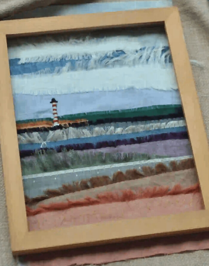A framed textile art piece depicting a landscape with a lighthouse, made out of things that would have been thrown away.