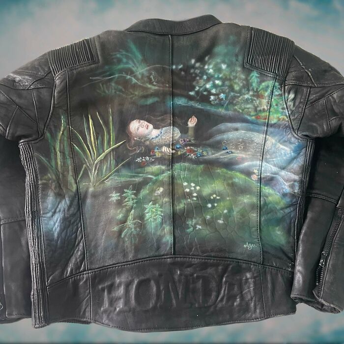 The back of a black leather jacket with an elaborate painting of a figure floating in water surrounded by greenery, made out of things that would have been thrown away.