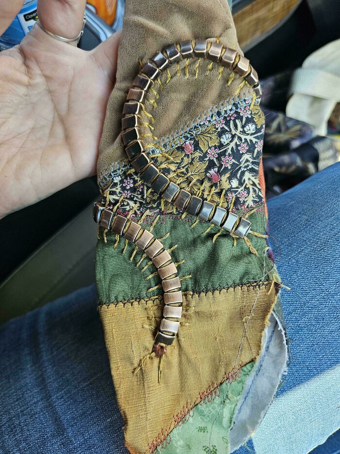 A person holding a decorative, patchwork fabric piece adorned with two curved rows of metal squares, made out of things that would have been thrown away.