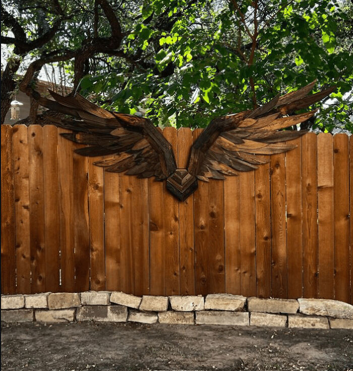 A pair of large, dark wooden wings attached to a rustic fence, made out of things that would've been thrown away.