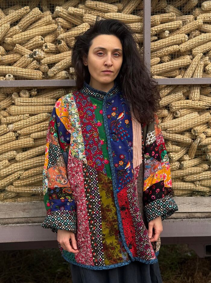 A woman wearing a colorful, patchwork coat made from things that would have been thrown away, standing in front of corn cobs.