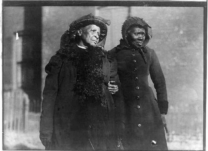 A historical photo showing two elderly women, one holding the other's arm, both dressed in dark coats and hats, feeling almost impossible to explain.