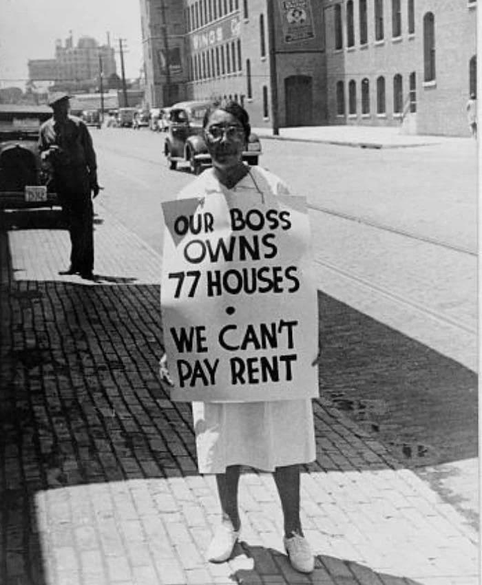 A historical photo of a woman holding a protest sign that reads Our boss owns 77 houses, we cant pay rent, feeling almost impossible to explain.