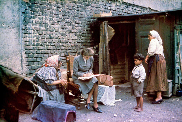 A historical photo showing a woman in a suit writing on a pad while two other women and a boy observe, feeling almost impossible to explain.