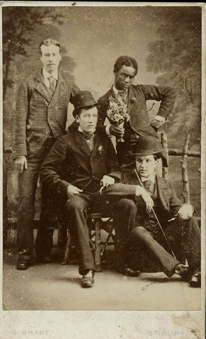 Four men in a historical photo, two standing and two sitting, wearing suits and hats, feeling almost impossible to explain.