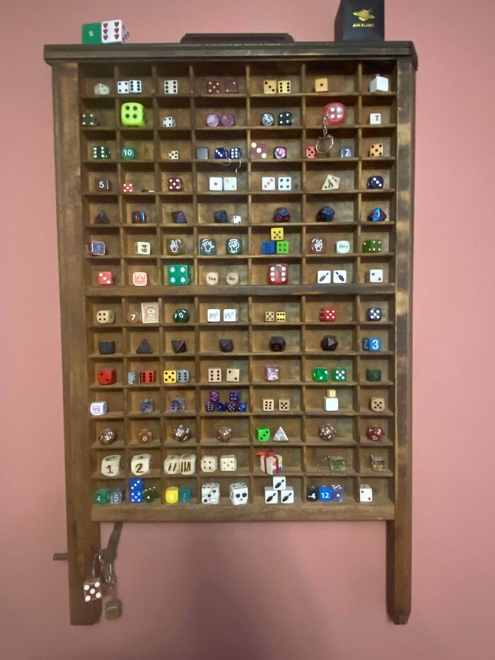 An unexpected and fascinating collection of various dice organized in a wooden display case hanging on a pink wall.