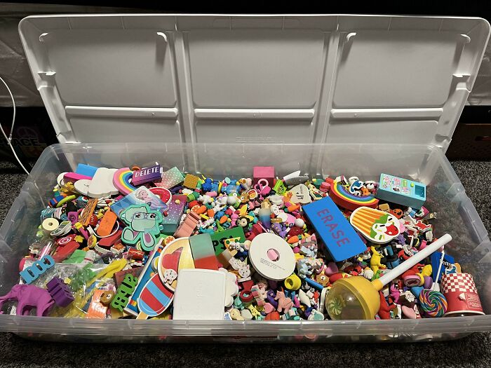 A large clear storage bin overflowing with an extensive collection of small, colorful erasers in various shapes.