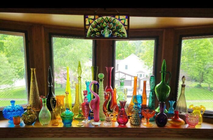 An array of vibrant glass bottles and vases, a fascinating collection, sits on a windowsill with a peacock stained glass.