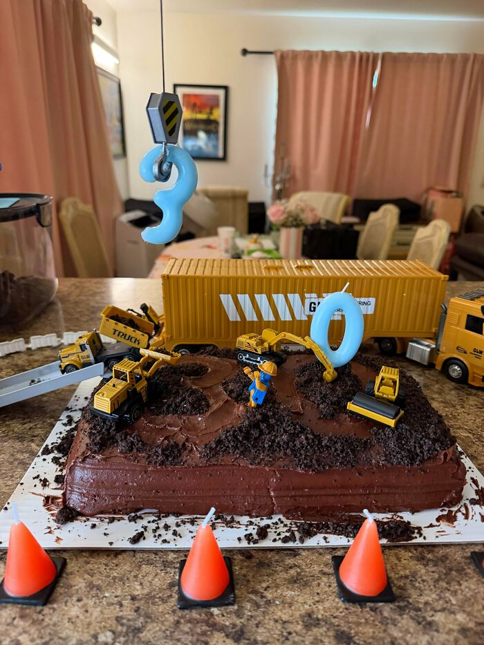 A construction-themed birthday cake for a 3-year-old, decorated with chocolate, toy trucks, and a crane hook, showing bakers amazing treats.