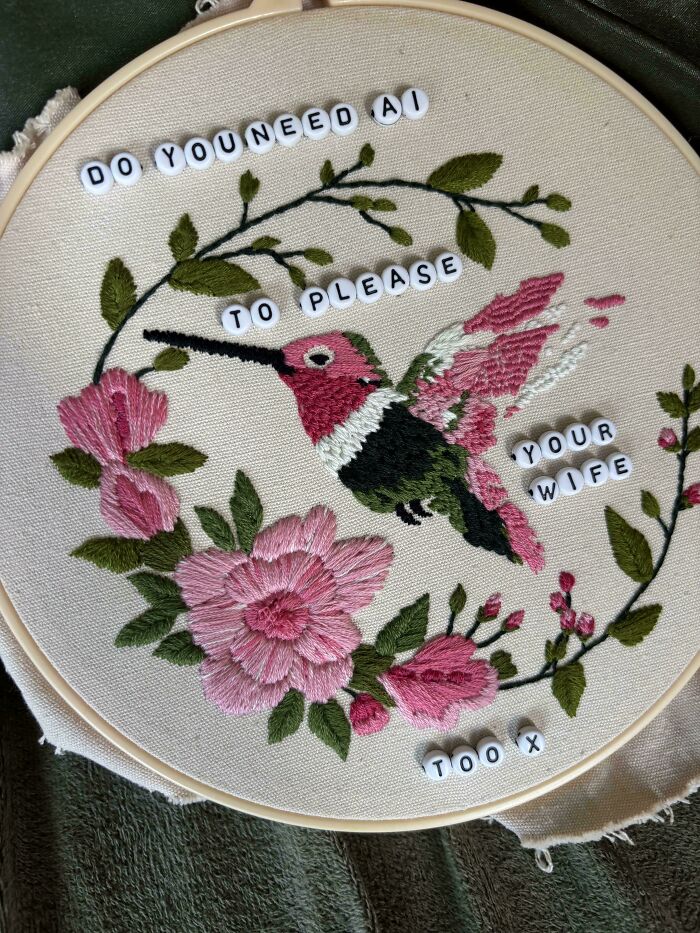 An embroidery piece depicting a pink hummingbird with flowers and bead text reading do you need AI to please your wife