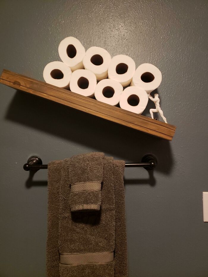 A humorous functional 3D print of a small figure holding up a wooden shelf with toilet paper rolls, making everyday life much easier.