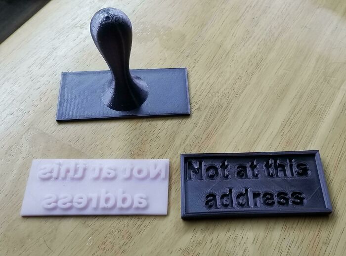 Two 3D printed rubber stamps and a stamp handle on a wooden table, featuring the phrase Not at this address for functional use.