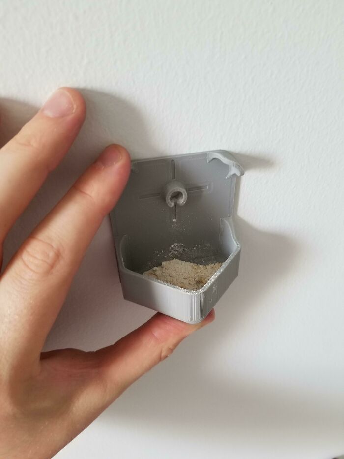 A hand holding a small gray 3D print attached to a wall, designed to make everyday life easier.