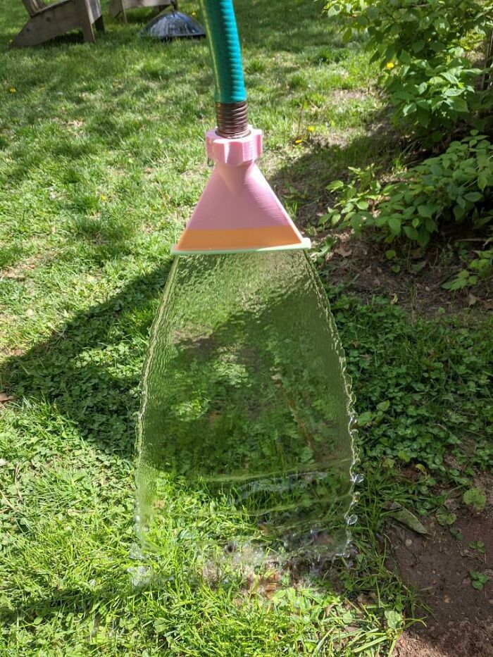 A pink and orange functional 3D print attached to a garden hose, spraying water in a wide arc.