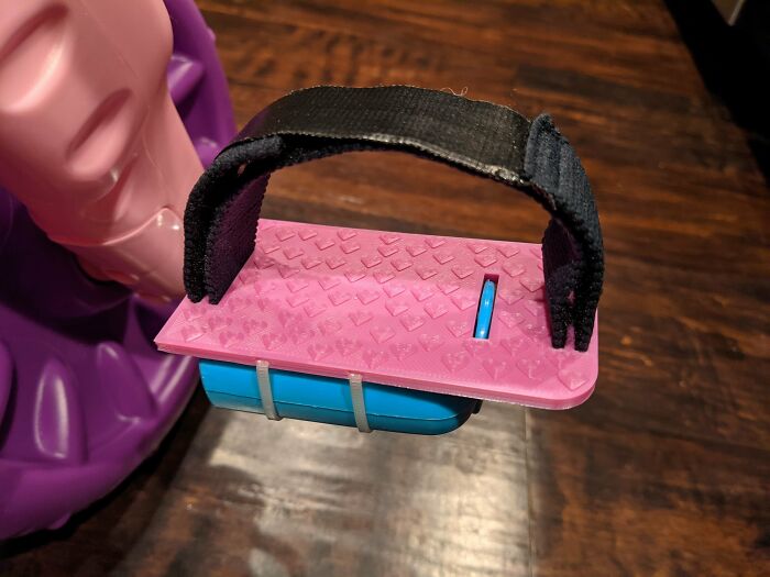 A functional 3D print: a pink and blue foot pedal attachment with a black strap, enhancing a child's ride-on.