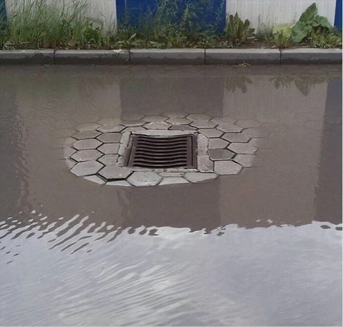 A storm drain surrounded by hexagonal pavers is submerged in standing water, illustrating a You Had One Job fail.